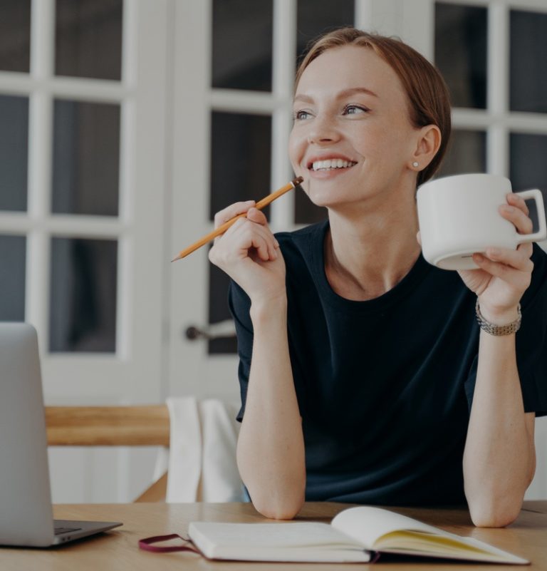 Smiling pensive woman holding pencil and cup write list in notebook, sitting at desk, looking to aside, dreaming, thinking. Happy dreamy female notes in notepad, planning day, visualizing good future.
